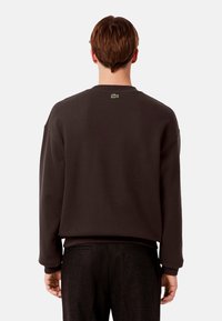 Sudadera marrón oscuro con cuello redondo, corte relajado, puños acanalados y un pequeño logo verde en la parte de atrás. Textura de tela suave.