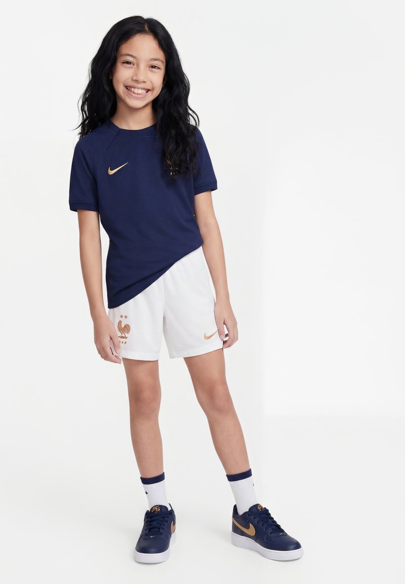 Fille souriante, debout avec une posture détendue, portant un t-shirt de sport bleu marine, un short blanc avec un logo, des chaussettes blanches et des baskets bleu marine avec des accents dorés.