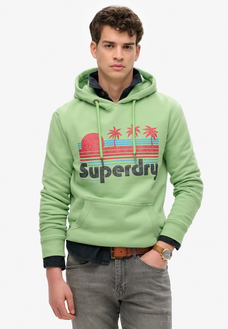 Sudadera verde claro con un estampado gráfico de un atardecer y palmeras en tonos rosa y azul, con el nombre de la marca "Superdry" en letras negritas negras.