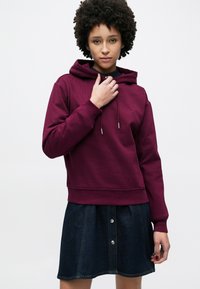 Felpa con cappuccio bordeaux con coulisse, tessuto strutturato e bordo a coste, indossata con una gonna di denim con bottoni decorativi e forma svasata.