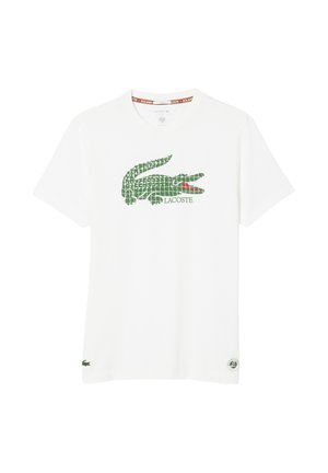 Camiseta blanca de manga corta con un gran logo del cocodrilo de Lacoste en verde y un pequeño cocodrilo verde cerca del dobladillo, con detalle rojo en la boca del logo.