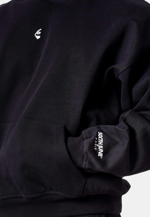 Zwarte sweatshirt met wit hartvormig logo op de borst en de tekst "SIXTH JUNE PARIS" op de manchet van de rechtermouw.
