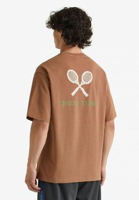 T-shirt marrone a maniche corte con grafica di racchette da tennis bianche e il testo verde "CIRCOLO TENNIS" sul retro. Realizzato in tessuto morbido.