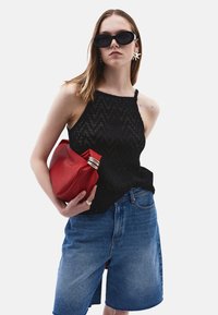 Top a fascia nera con texture, pantaloni corti in denim blu, pochette rossa con accenti metallici, occhiali da sole ovali neri, modella con capelli lisci.