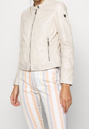 Mujer con chaqueta de cuero cremosa con cremallera y cuello con botón a presión, y pantalones pastel a rayas, de pie frente a un fondo liso.