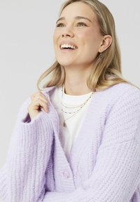 Cardigan en tricot lavande avec fermeture à boutons, texture côtelée, manches larges, porté par-dessus une chemise blanche, superposé avec des bijoux délicats.