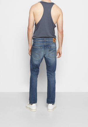 Homme portant un jean bleu, des baskets blanches et un débardeur racerback gris foncé, debout face à un mur blanc.