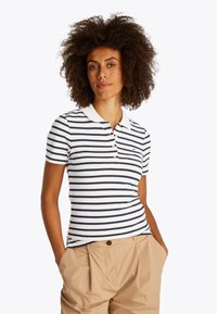 Kobieta o kręconych włosach, ubrana w biało-granatową koszulkę polo w paski i beżowe spodnie, z rękami w kieszeniach na białym tle.