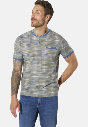 Mann steht, trägt ein kurzärmeliges gestreiftes Hemd mit blauem Kragen und Jeans, linker Arm tätowiert, schaut leicht nach links.