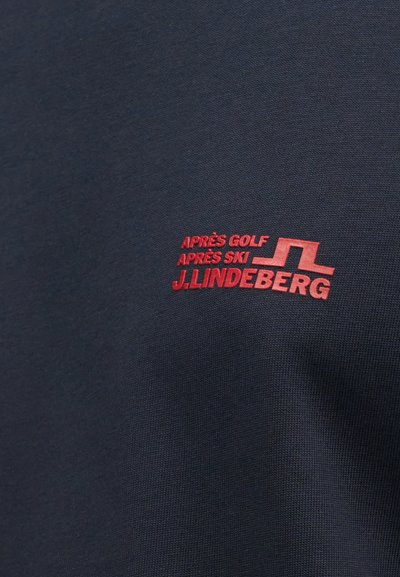Piros "APRÈS GOLF APRÈS SKI J. LINDEBERG" logó sötét tengerészkék anyagon, nagybetűs hímzéssel és stilizált emblémával.