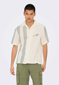 Camisa de manga corta en color crema con un patrón de cuadros negros en un lado y detalle bordado que dice 'Capri'. Tejido ligero, diseño con cuello.