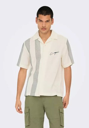 Camisa de manga corta en color crema con un patrón de cuadros negros en un lado y detalle bordado que dice 'Capri'. Tejido ligero, diseño con cuello.
