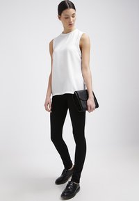 Top blanco sin mangas con tejido texturizado, combinado con pantalones negros ajustados y zapatos negros, sosteniendo un bolso de mano negro. Diseño sencillo.