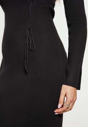 Schwarzes, geripptes Strickkleid mit taillierter Form, seitlichen Zügen und langen Ärmeln, präsentiert mit einer Hand, die einen goldenen Ring trägt.