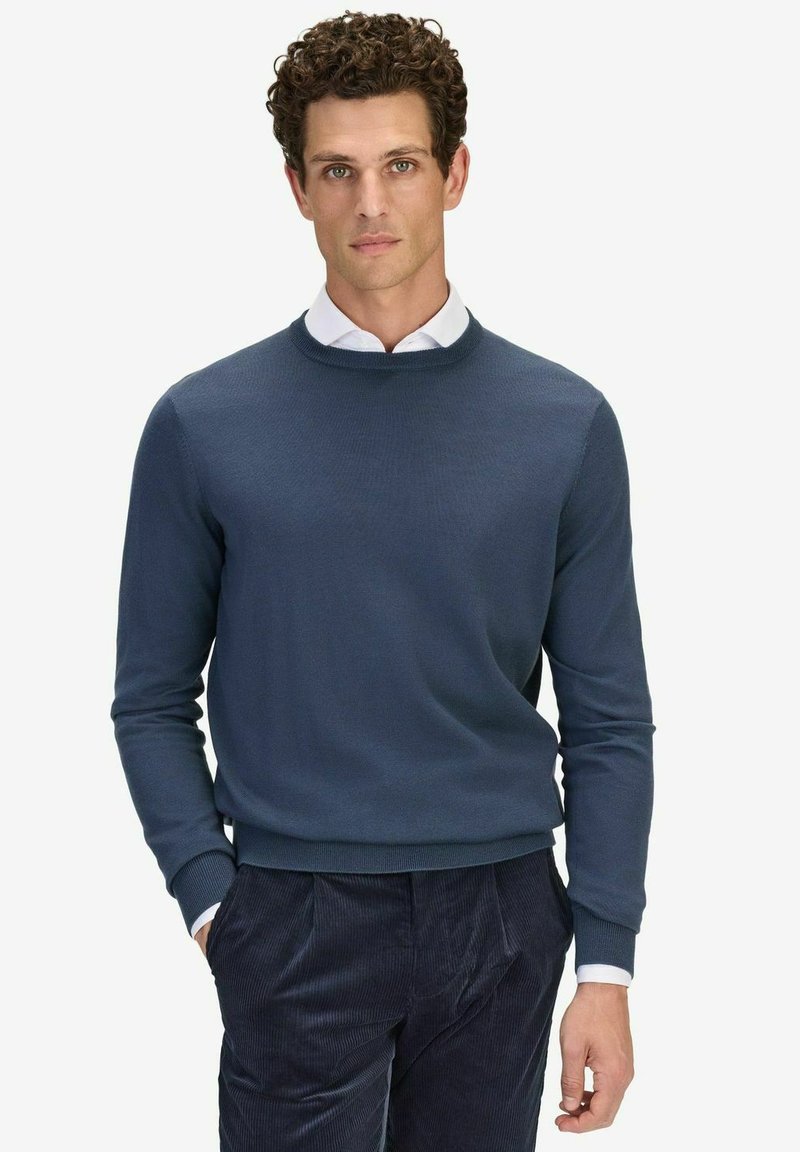 Mörkblå crewneck tröja i stickad tyg, med ribbade manschetter och nederkant, i kombination med mörka manchesterbyxor, bärs över en vit skjorta.