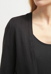 Kvinna iklädd en svart cardigan över en svart top med rundad hals, visad från nacke till övre bröst och axlar framför en neutral bakgrund.