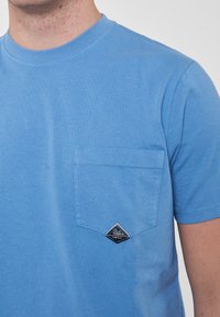 T-shirt di cotone blu con maniche corte, collo rotondo e tasca frontale con un'etichetta a forma di diamante. Texture morbida, vestibilità regolare.