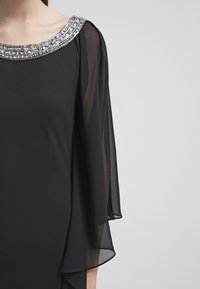 Robe noire avec un col orné de bijoux, des manches transparentes et une silhouette fluide. Le tissu est lisse avec un léger éclat.