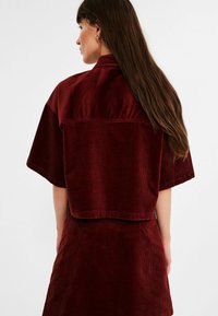 Burgundy corduroy crop-top with short sleeves, featuring a boxy cut and visible ribbed texture, paired with a matching skirt.