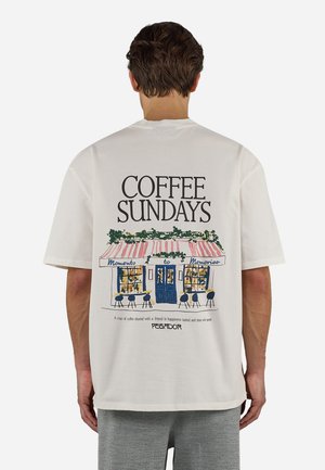 Άνδρας που φοράει λευκό μπλουζάκι με εικονογράφηση καφετέριας, κείμενο "COFFEE SUNDAYS" και φράση για τον καφέ και τη φιλία στην πλάτη.