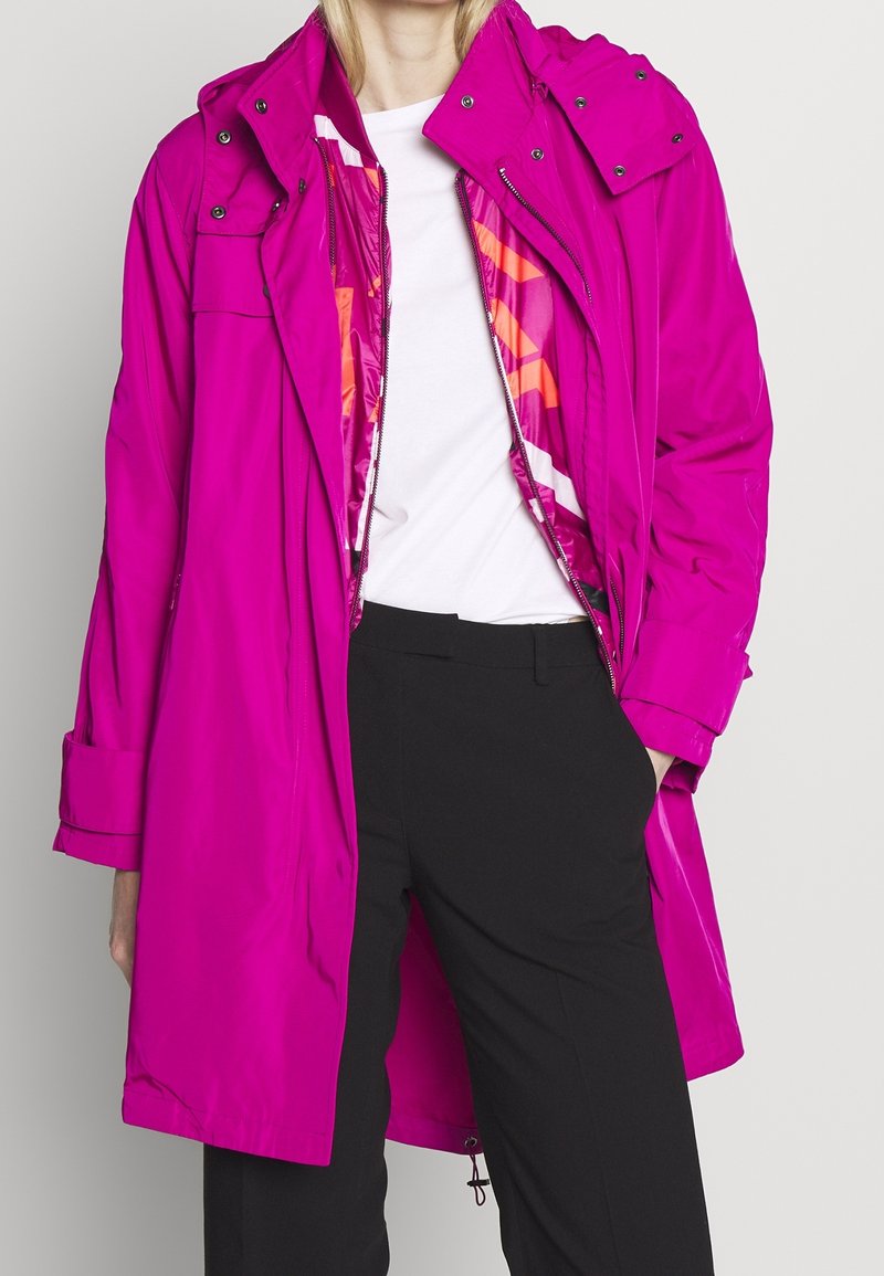 Roze waterdichte regenjas met grote capuchon, ritssluiting aan de voorkant en zakken. Lichtgewicht stof met een gladde textuur en een kleurrijke voering aan de binnenkant.