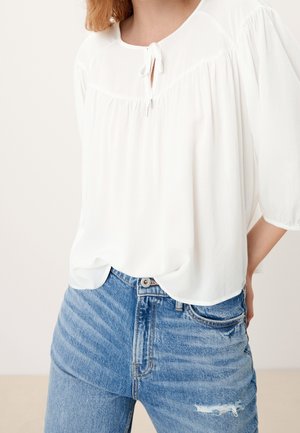 Femme portant un chemisier blanc ample avec un lien au niveau du col et un jean bleu déchiré taille haute avec fermeture par boutons.