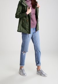 Veste à capuche vert olive, superposée sur un pull violet, associée à un jean bleu clair déchiré et des baskets argentées.