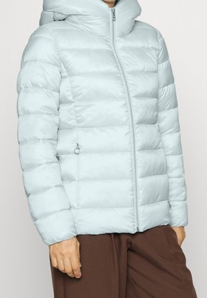 Lyseblå quiltet dunjakke med høj krave og lynlås foran, båret af en person iført brune bukser.