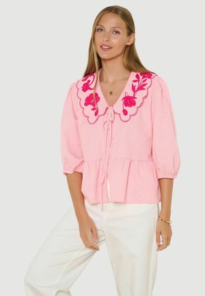 Vrouw met lang blond haar, gekleed in een roze blouse met pofmouwen en een bloemenborduurwerk op de kraag, gecombineerd met een witte broek.