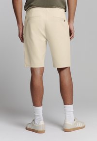 Beige shorts met een gladde textuur, twee achterzakken en een middellange snit. Het model draagt witte sokken en beige sneakers.