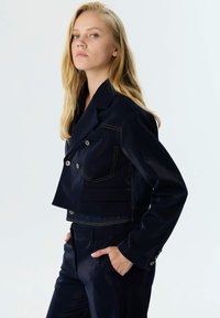 Marineblauwe cropped jas met een dubbelzijdig ontwerp, voorzien van contrasterende oranje stiksels, twee voorzakken en metalen knopen.