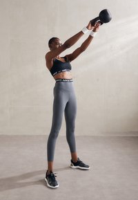 Svart sportsbh med hvit logo, grå leggings, svarte treningssko, og hvite håndleddsbånd. Holder en svart kettlebell over hodet.