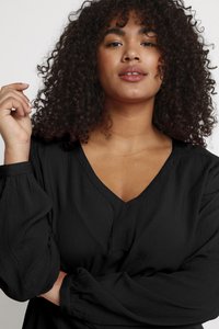 Blouse noire à col en V avec manches longues bouffantes. Le tissu a une finition texturée et est drapé de manière lâche autour du corps.