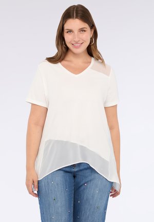 Mujer con cabello castaño que lleva pendientes de aro, una blusa blanca de manga corta y escote en V con dobladillo transparente, y jeans azules con pequeños adornos coloridos.