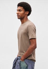 Νέος άνδρας με αφρο μαλλί, φορώντας μπεζ κοντομάνικη μπλούζα, ανοιχτόχρωμο μπλε τζιν και μπλε καρό πουκάμισο δεμένο γύρω από τη μέση του.