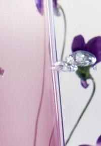 Verre rose transparent avec une surface lisse, présentant des gouttes d'eau et des accents floraux violets en flou artistique.