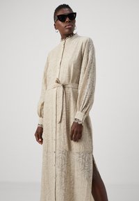 Long beige dress with a leaf pattern, button-down front, tie belt, and cuffed sleeves featuring lace trim. Smooth texture with side slits.