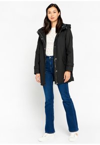 Zwarte waterdichte parka-jack met een capuchon, voorzien van zilverkleurige hardware en zijzakken, gedragen over een witte gebreide top en blauwe flare jeans.