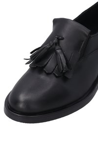Černá kožená slip-on obuv s texturovaným povrchem a ozdobnými černými třásněmi na nártu. Zaoblená špička a plochá podrážka.