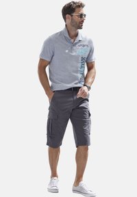 Graues Poloshirt mit bedrucktem Textdesign, kombiniert mit dunklen Cargoshorts und weißen Sneakers. Modell trägt Sonnenbrille und steht mit den Händen in den Taschen.