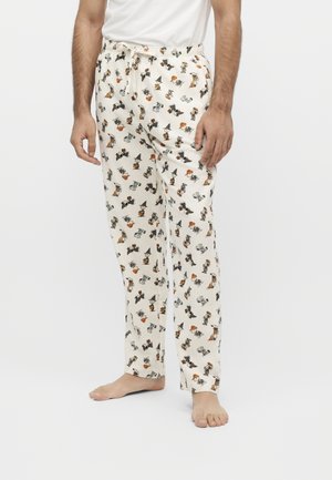 Homme portant un pyjama blanc avec de petits personnages de dessin animé d'Halloween éparpillés, debout pieds nus devant un fond uni.