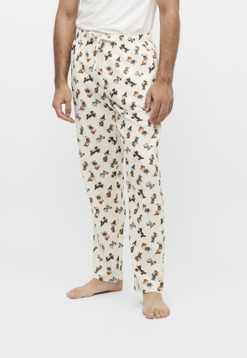 Homme portant un pyjama blanc avec de petits personnages de dessin animé d'Halloween éparpillés, debout pieds nus devant un fond uni.