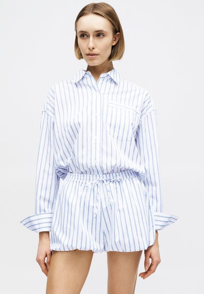 Γυναίκα που φορά λευκό πουκάμισο μακρυμάνικο και ταιριαστά σορτς με κάθετες μπλε ρίγες, στέκεται μπροστά από ένα απλό φωτεινό φόντο.