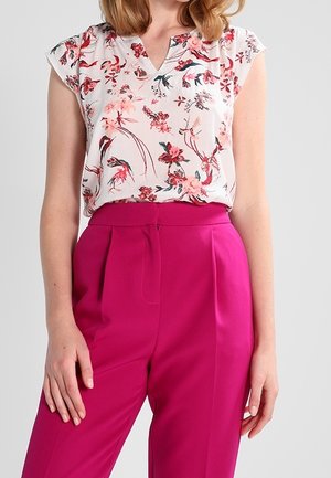 Femme portant un chemisier blanc à manches courtes avec un imprimé floral rose, rentré dans un pantalon taille haute rose vif.
