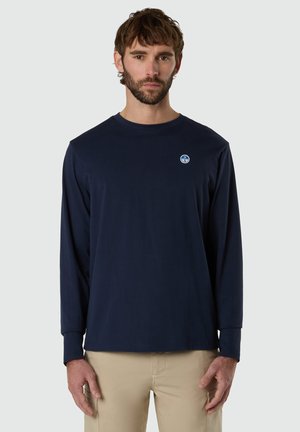 Mann mit kurzen braunen Haaren und Bart, der ein marineblaues Langarmhemd mit einem kleinen runden Logo auf der Brust und beige Hosen trägt.