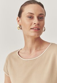 Top beige con scollo rotondo e bordi bianchi, realizzato in tessuto morbido. Il modello indossa orecchini a cerchio dorati su uno sfondo neutro.