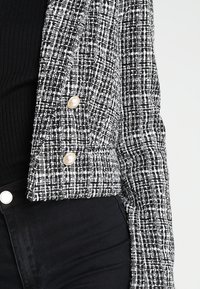 Blazer en tweed noir et blanc avec un motif texturé, doté de boutons en perle, coupe ajustée et col à revers, porté sur un haut noir.