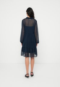 Φόρεμα από διάφανο ύφασμα σε ναυτικό μπλε, με μακριά μανίκια, φούστα με στρώσεις και εφαρμοστό μπούστο με τετράγωνη πλάτη. Διακοσμημένο με απλές ραφές.