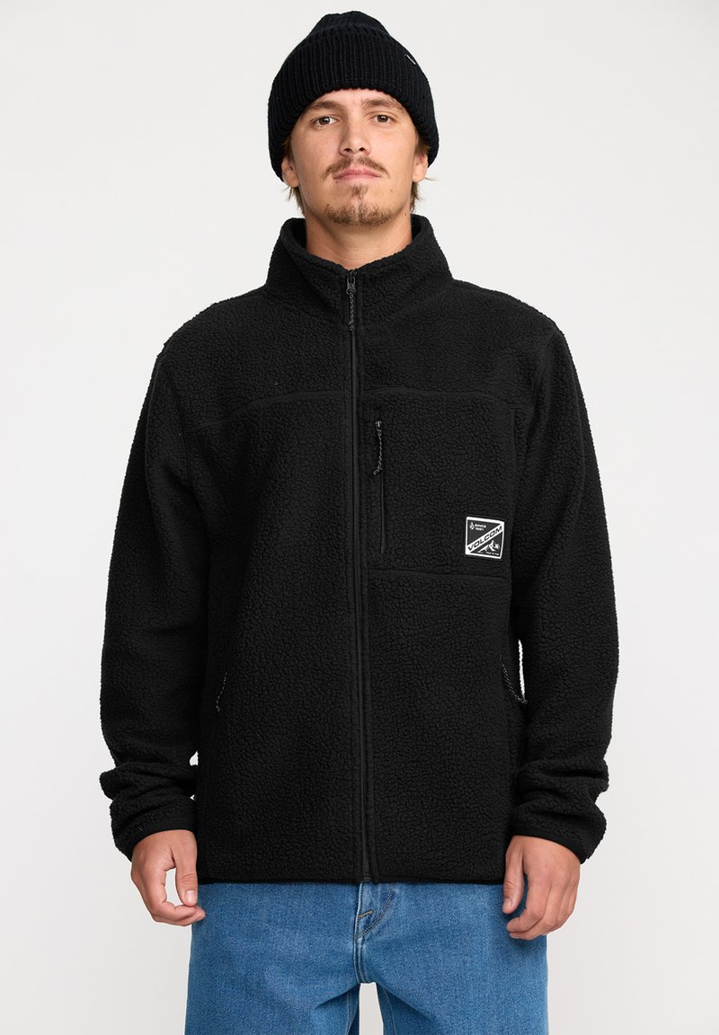 Svart fleecjacka med dragkedja fram, hög krage och två fickor. Har en logopatch på vänster bröst. Bärs över blå denim.