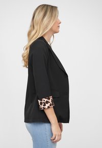 Svart blazer med strukturert design og trekvartlange ermer, med leopardmønster på mansjettene. Brukt over lyseblå jeans.